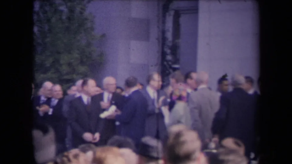 Ceremony attendees in suits and coats gather near a doorway and trees while the crowd applauds in soft 8mm color.