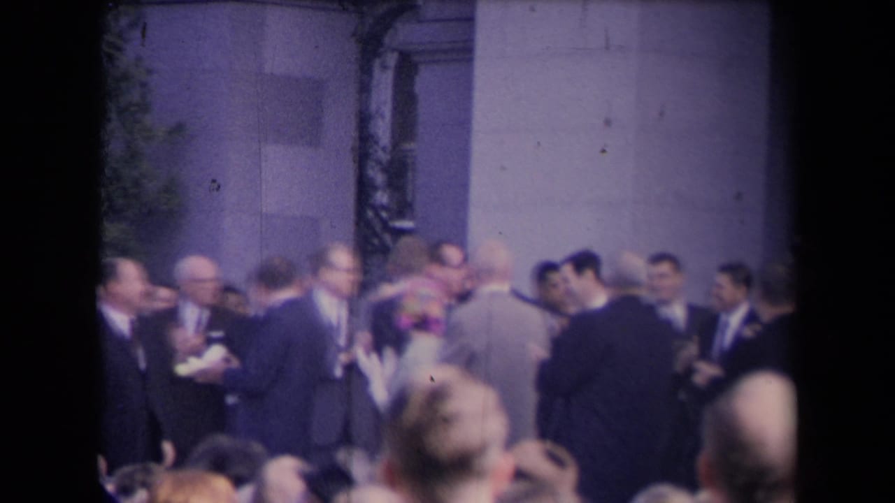 Soft 8mm frame of suited men and spectators standing near a stone building during a formal public gathering.