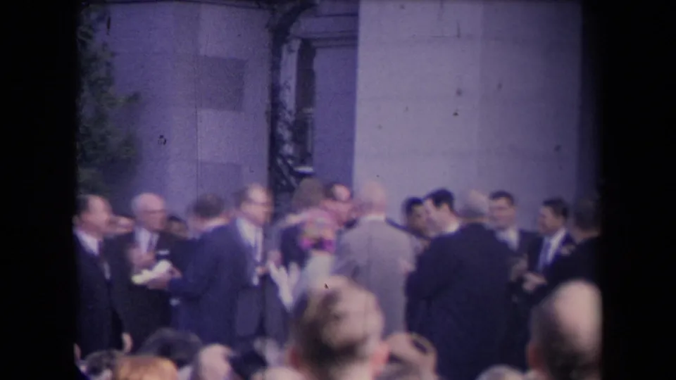 Soft 8mm frame of suited men and spectators standing near a stone building during a formal public gathering.