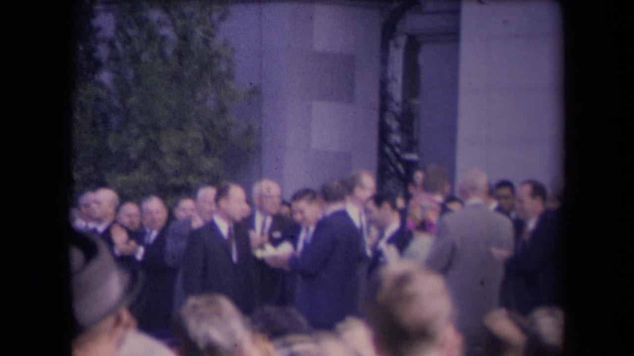 Formally dressed men, some applauding, stand among a dense crowd outside a stone building in archival 8mm footage.