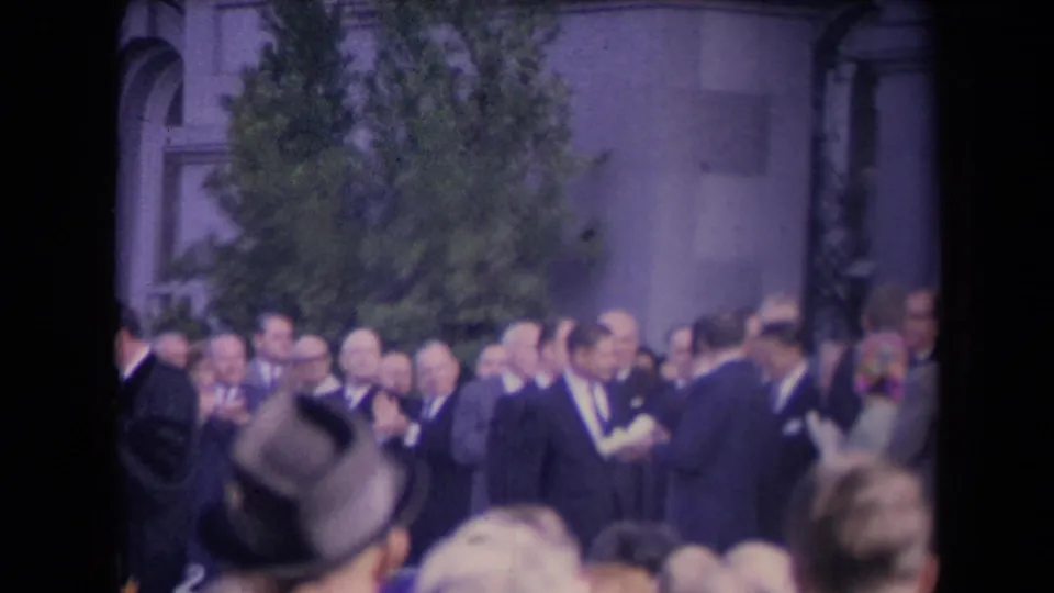 Handheld 8mm view across spectators toward suited men gathered near a tree and formal stone architecture.