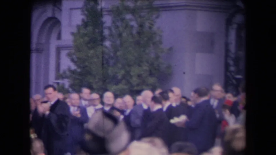 Crowded 8mm frame showing suited attendees, foreground spectators, a tree, and stone architecture during a formal public event.