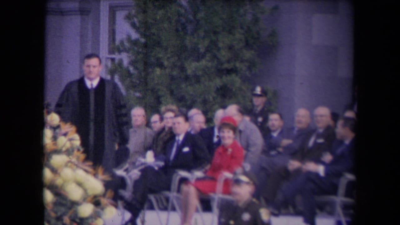 Robed minister walks near seated adults during an outdoor formal ceremony.