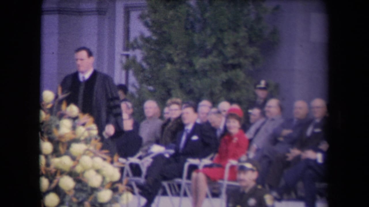 The minister moves past pale flowers while men in suits and a woman in red sit in rows at a formal outdoor ceremony.