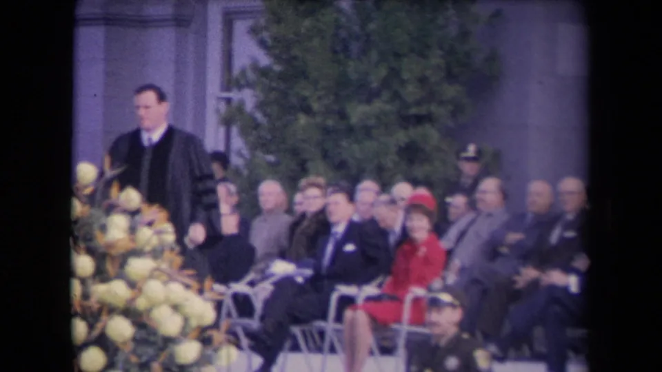The minister moves past pale flowers while men in suits and a woman in red sit in rows at a formal outdoor ceremony.