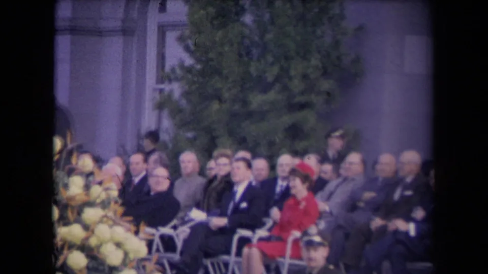 A formally dressed audience sits outdoors near a large flower arrangement and building facade in soft 8mm color footage.