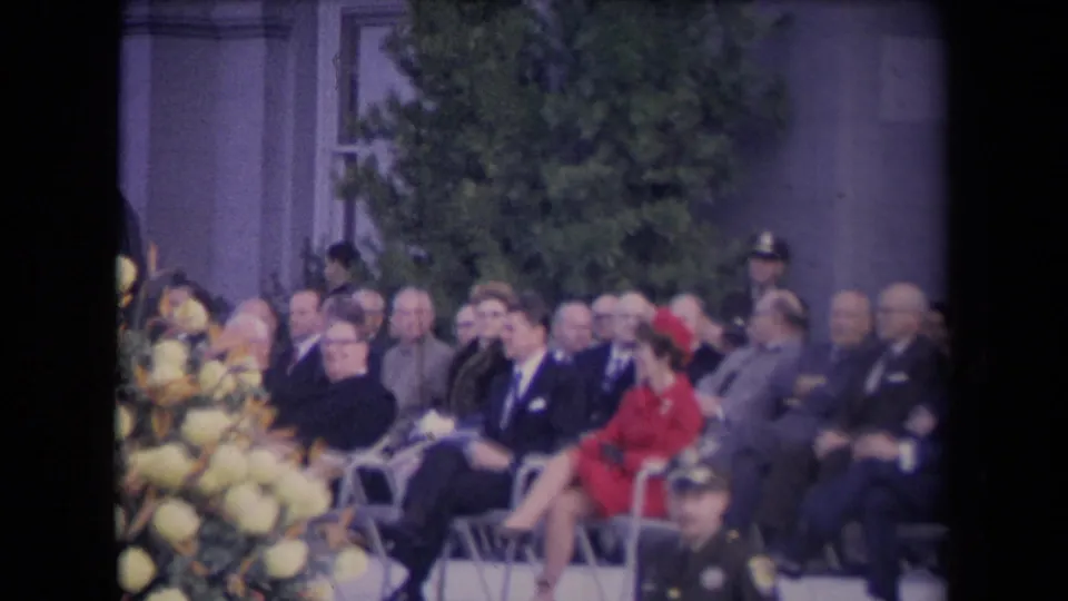Rows of formally dressed adults sit beside pale flowers, with a woman in red and uniformed personnel visible in the 8mm frame.