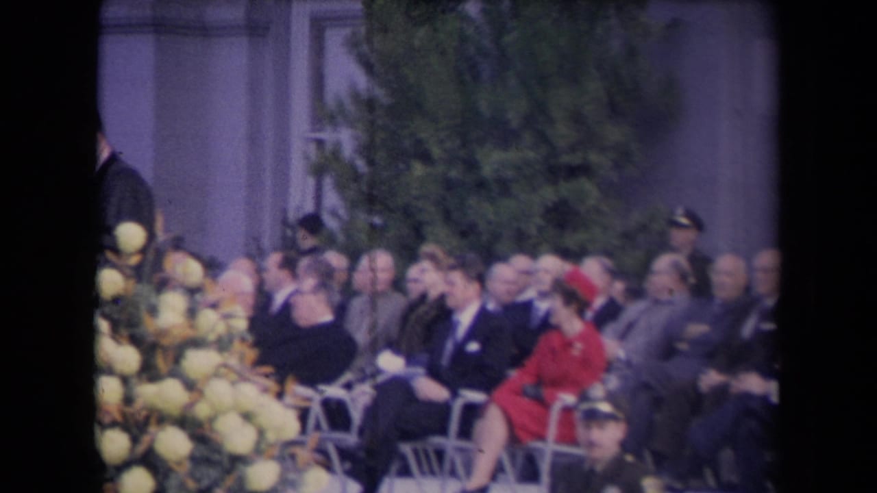 Seated audience at a formal outdoor ceremony with flowers in the foreground.
