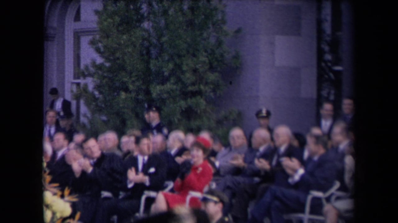 Formally dressed adults applaud in outdoor seating, with a woman in red visible among the crowd and uniformed personnel behind them.