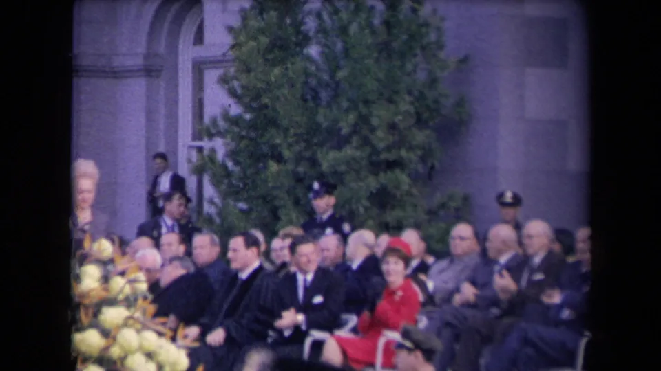 Outdoor ceremony audience with men in dark suits, a woman in a red outfit, floral display, and uniformed personnel near the building.