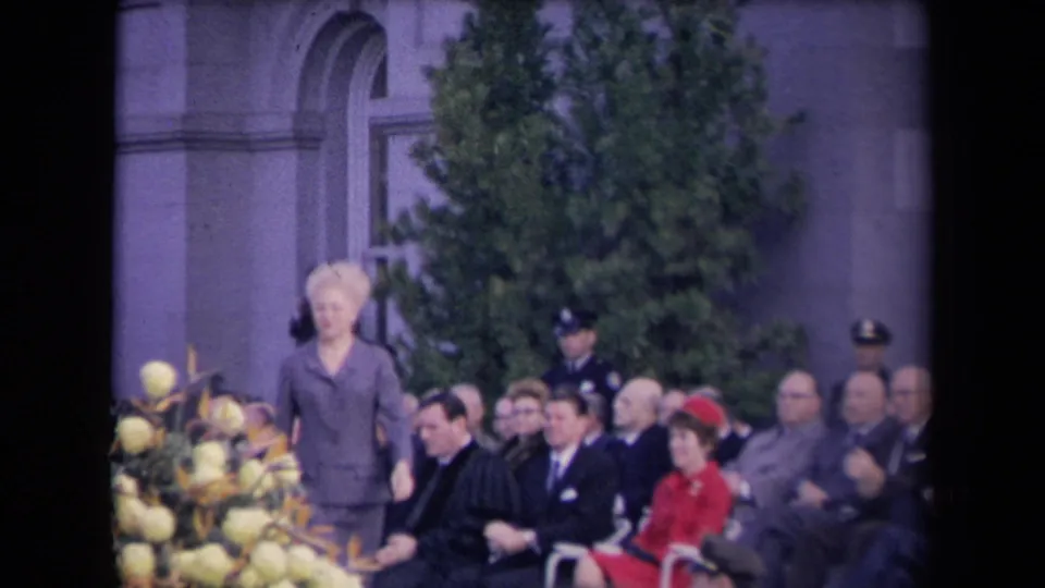 A woman in a gray suit stands near a floral arrangement while seated guests, including a woman in red, look on and applaud.