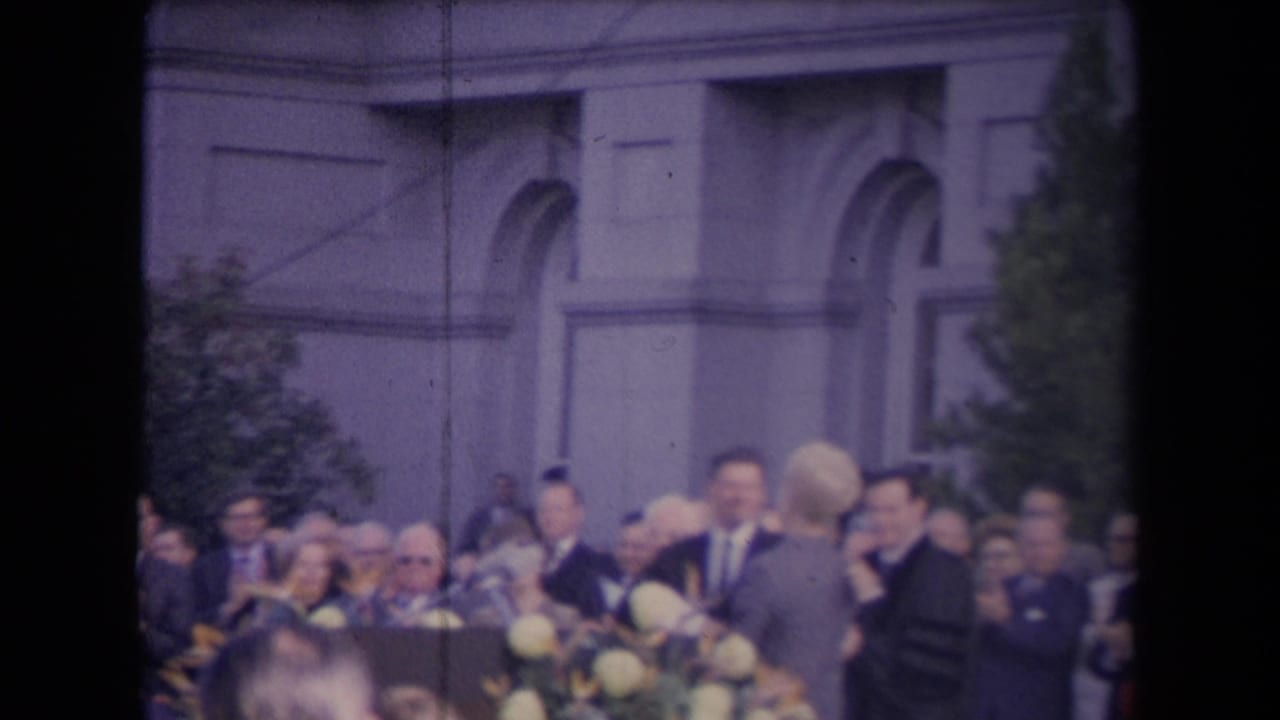 Attendees face a podium area with microphones and pale flowers in front of a stone building facade.