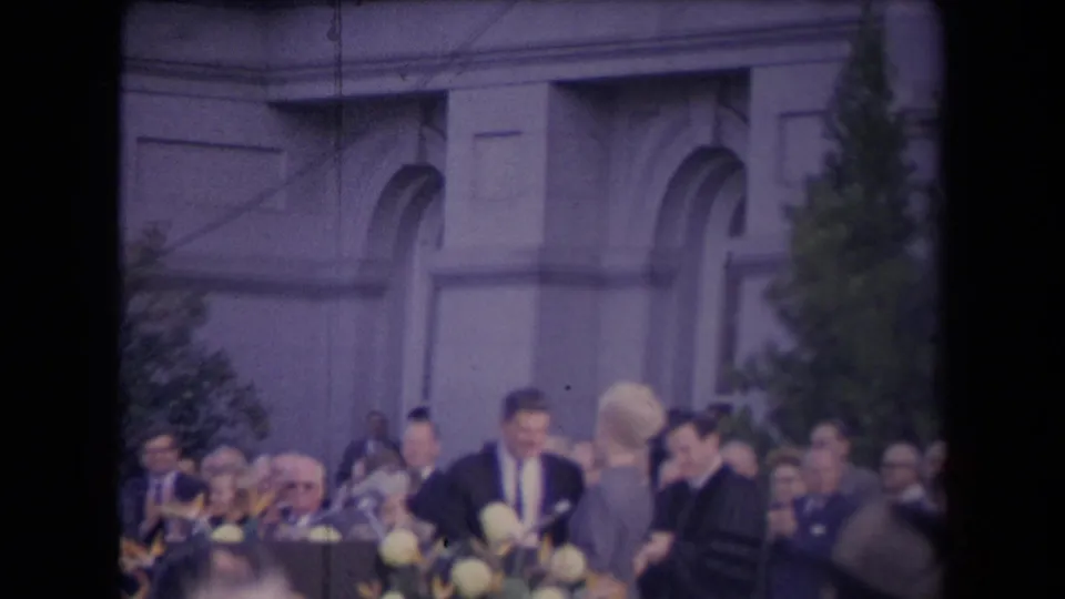 A suited man near the microphone area faces a woman in front of a seated crowd and formal building facade.