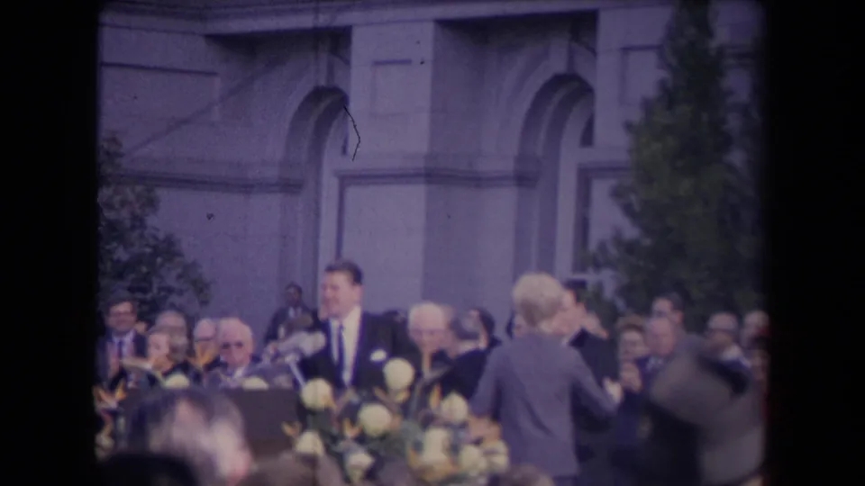 A suited speaker stands behind microphones and flowers while attendees watch and applaud outdoors.