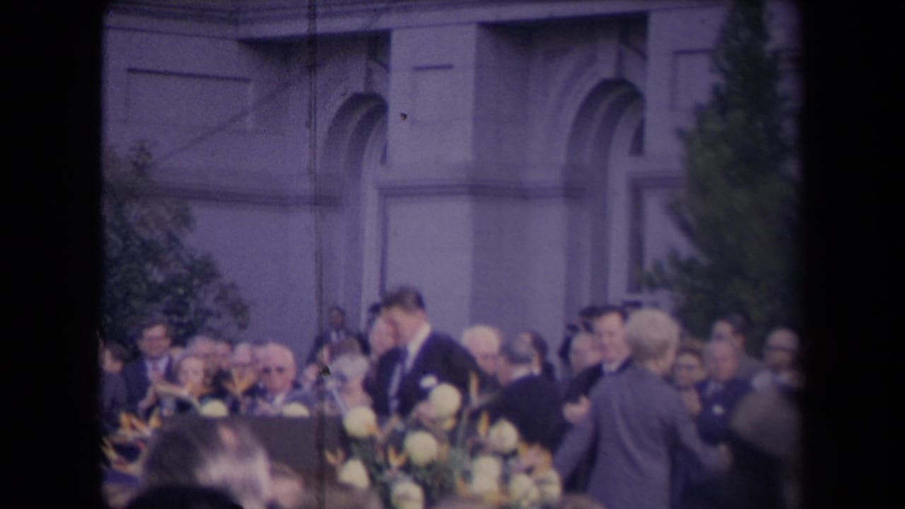 1967 8mm frame of formally dressed adults applauding at an outdoor ceremony, with flowers and a classical building facade visible.