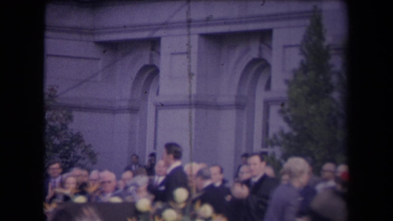 Outdoor 1967 ceremony frame showing a suited man and surrounding crowd near floral arrangements before an arched building facade.