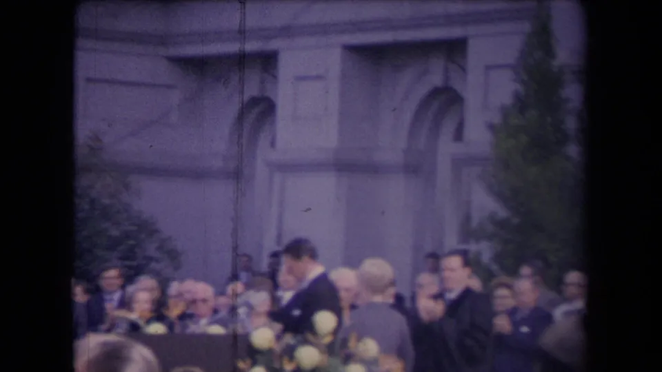Soft 8mm frame with the ceremony partly blocked by foreground heads, showing adults, flowers, and a civic building backdrop.