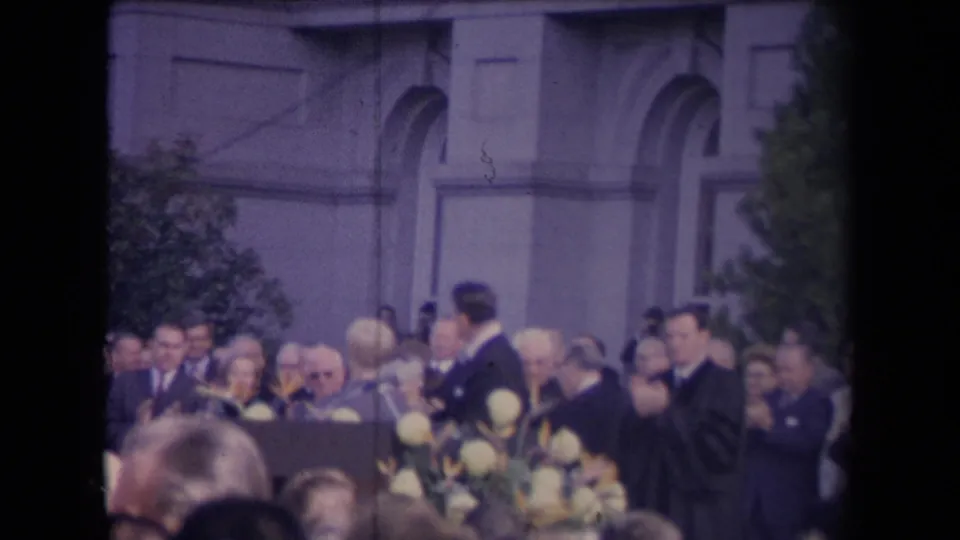 Outdoor 1967 inauguration footage frame showing adults applauding around a suited figure, with flowers and a large facade behind them.
