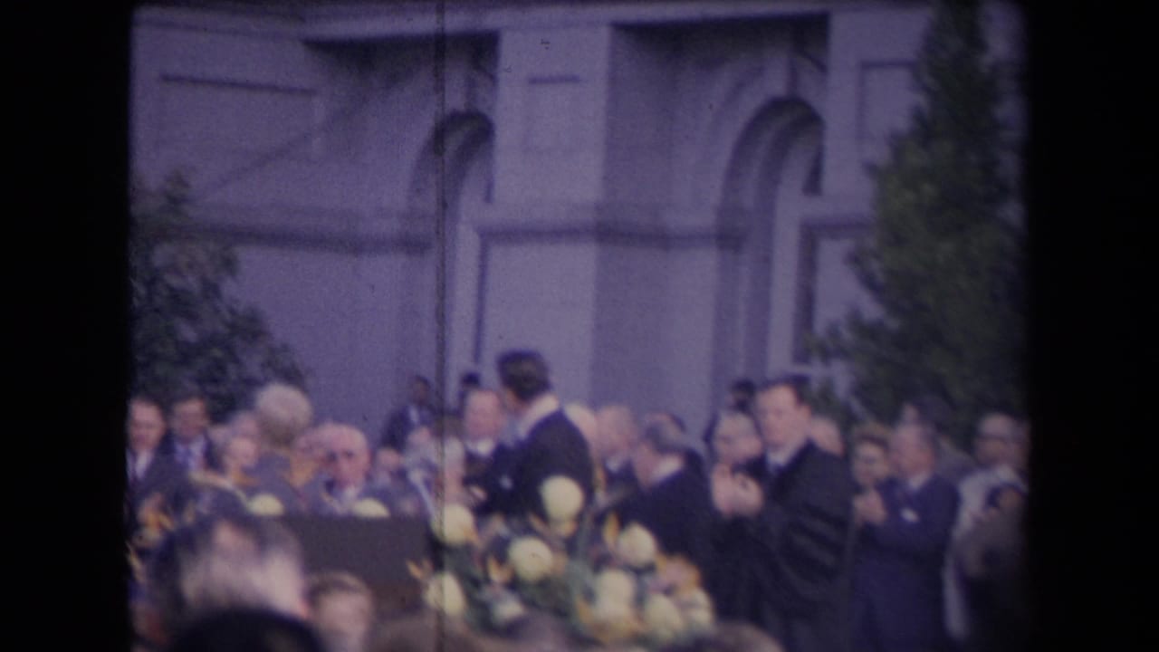 A suited man stands near a podium with white flowers as attendees sit outside a formal building, supported by 1967 political-event metadata.