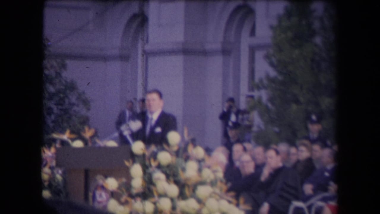 A suited speaker addresses an outdoor audience from a flowered podium, with photographers and arched windows visible in the background.