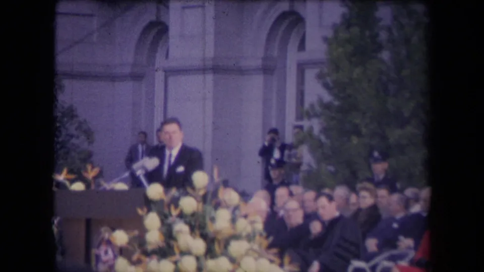 A suited man stands behind a podium at an outdoor ceremony, framed by flowers, seated attendees, trees, and a formal building facade.