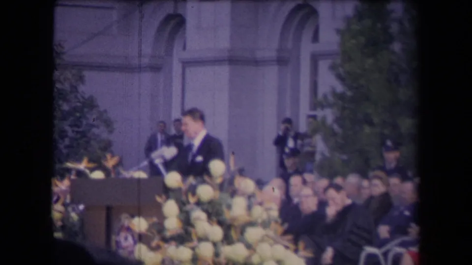 The suited speaker turns slightly at a flowered podium as seated attendees watch, with photographers, trees, and arched windows in the background.