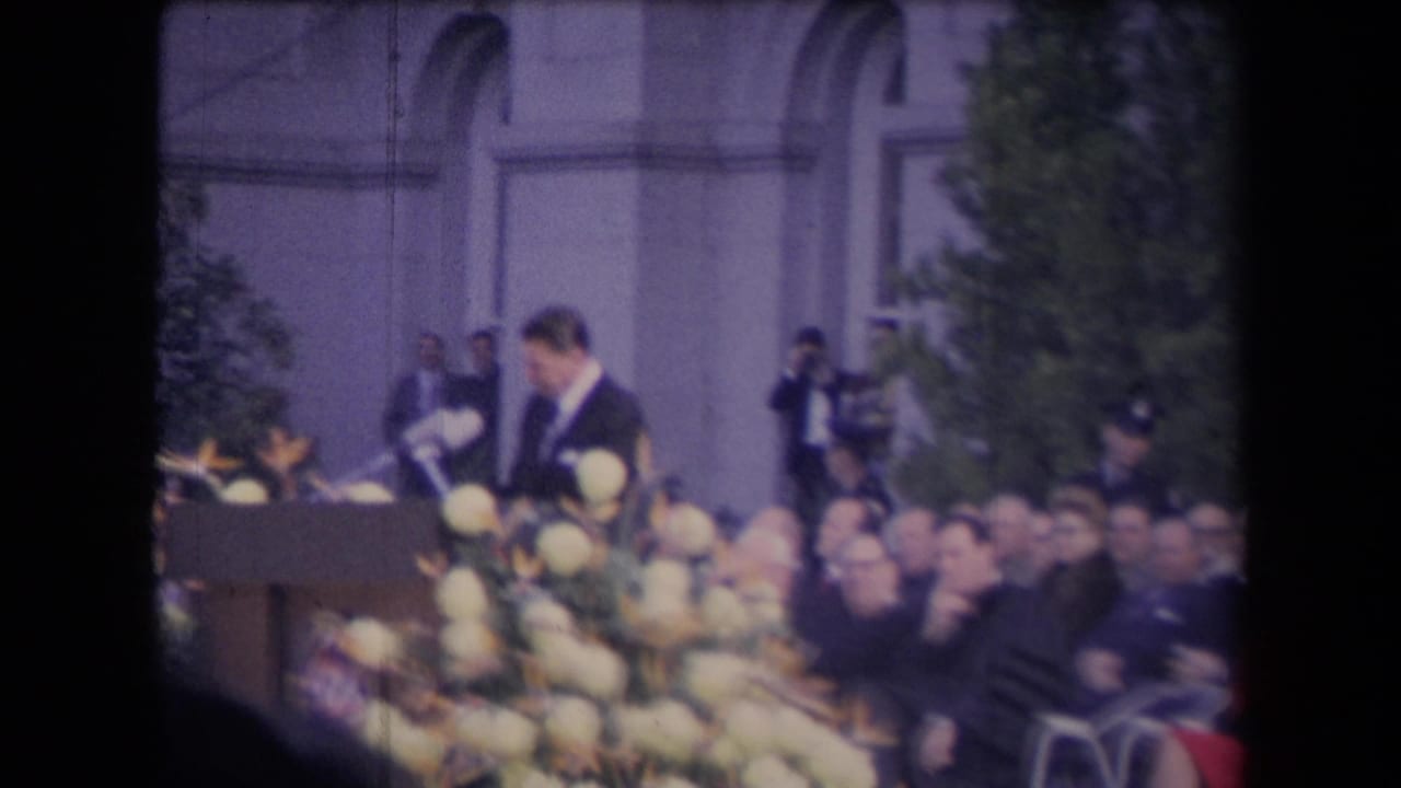 The speaker remains at the podium as the camera frames flowers, seated attendees, and staff or security near the building behind him.