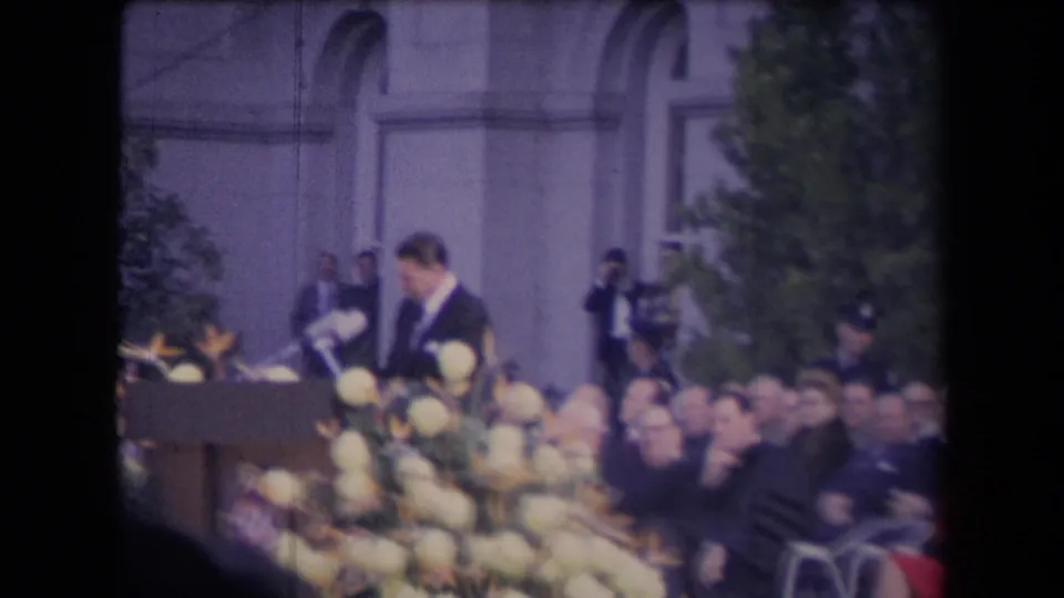 The speaker remains at the podium as the camera frames flowers, seated attendees, and staff or security near the building behind him.