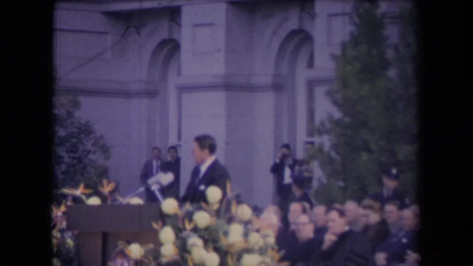 Reagan appears turned away from the podium during the outdoor ceremony, with flowers, seated spectators, and onlookers near the building.