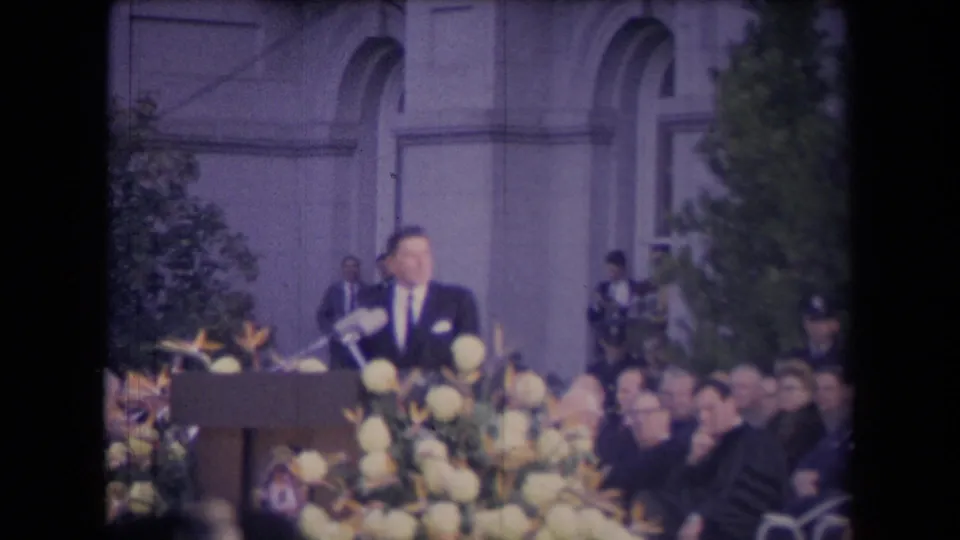 Reagan faces the crowd from the podium, framed by flower arrangements, seated attendees, trees, and the institutional building behind him.