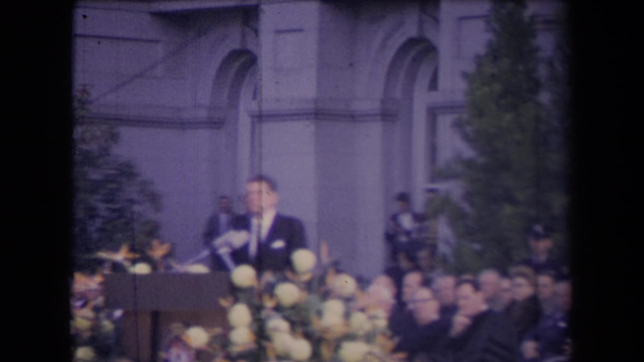 Suited man speaking at a flower-lined podium before a seated audience outside a large building.