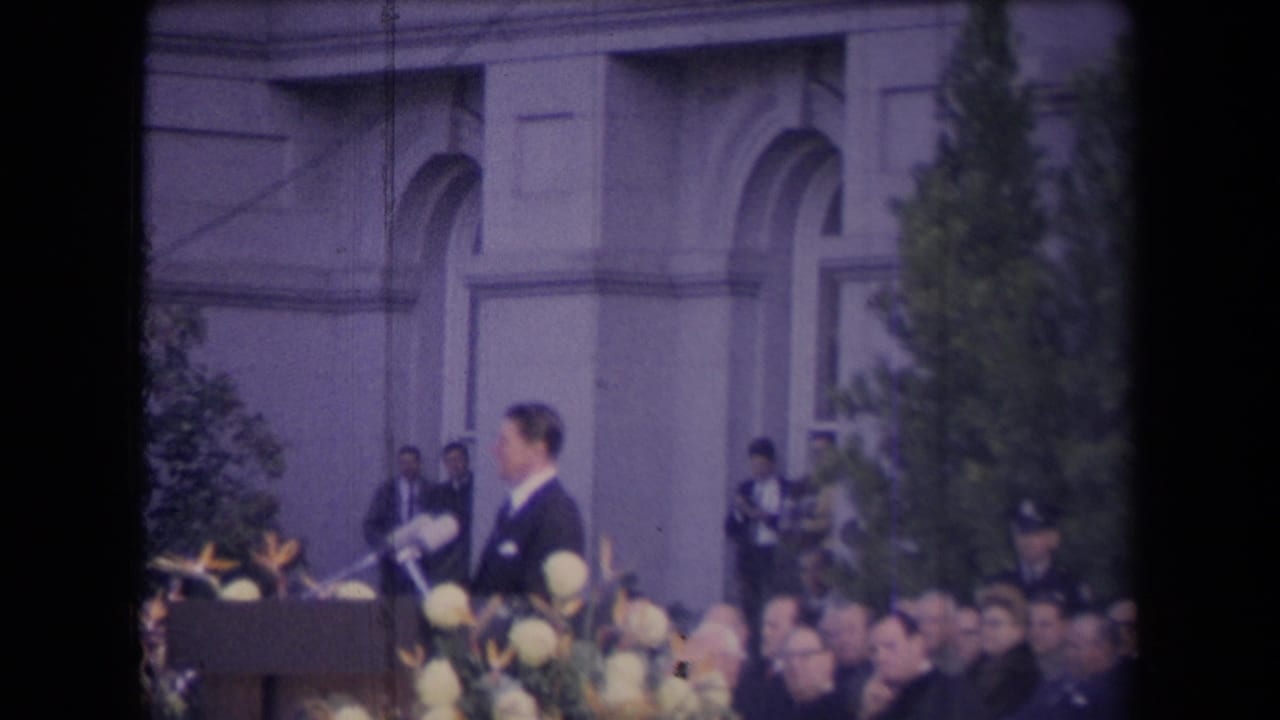 The speaker turns slightly at the podium during the documented 1967 ceremony, with microphones, flowers, audience, trees, and security personnel in view.