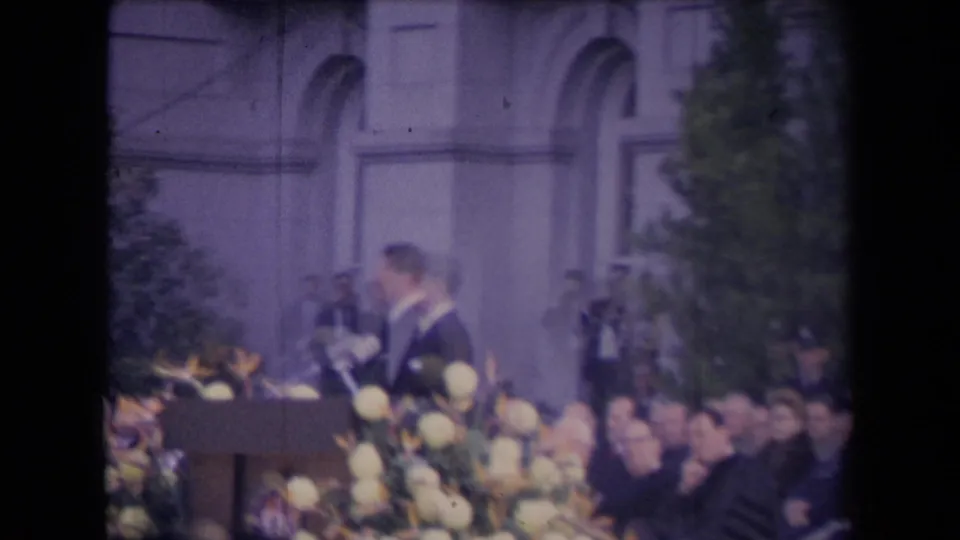 A soft handheld frame shows the suited speaker in profile near the podium, with floral decorations, seated spectators, and the building facade behind him.
