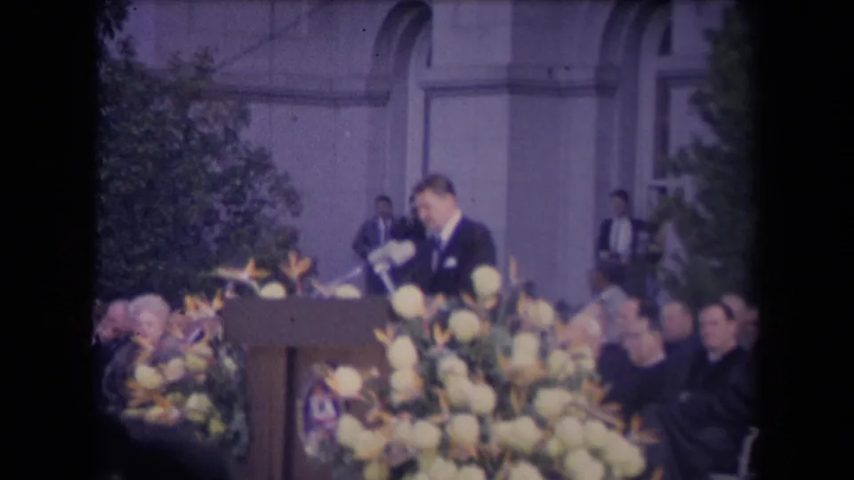 The speaker leans toward the microphone cluster at the decorated podium, framed by flowers, audience members, trees, and the formal building exterior.