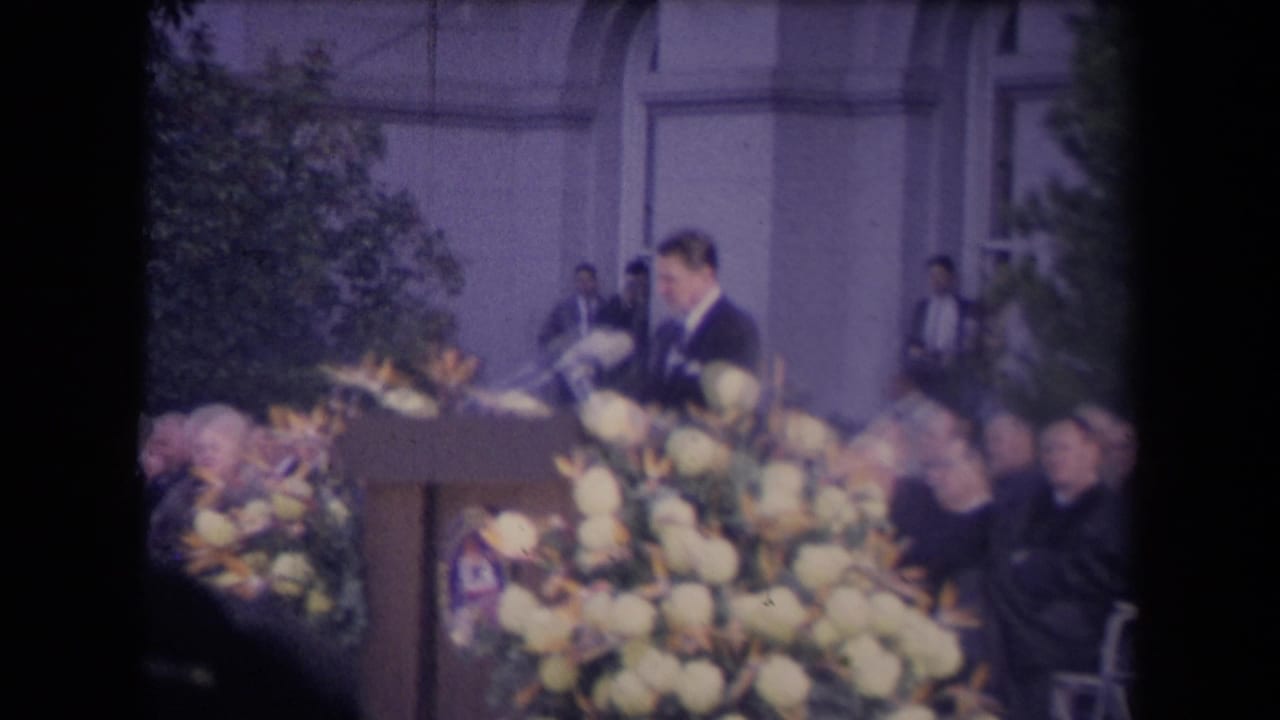 Tall man in a dark suit speaks at a flower-covered podium before seated guests.