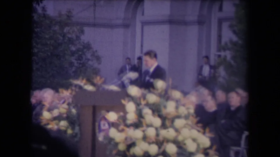 A tall suited speaker stands at a wooden podium with microphones and large flowers during a formal 1960s civic ceremony.