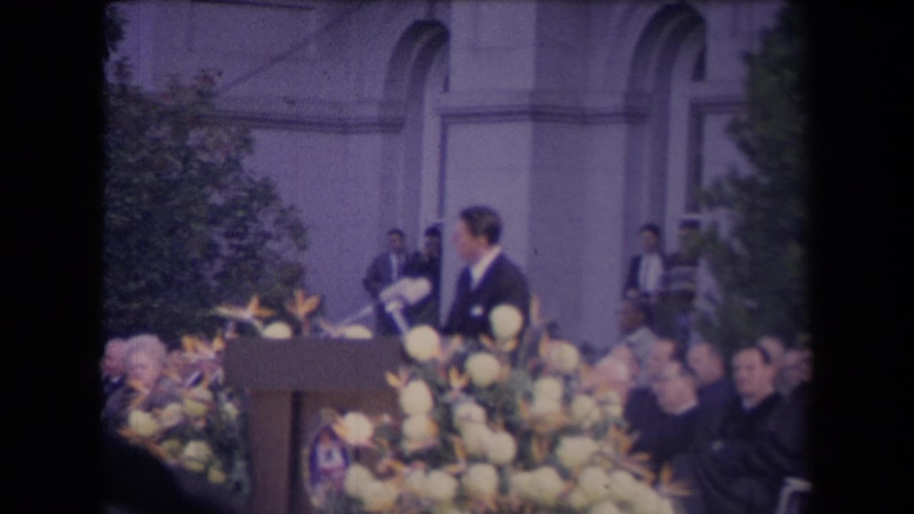 The speaker is seen in profile at the floral podium, with audience members and arched windows behind him.