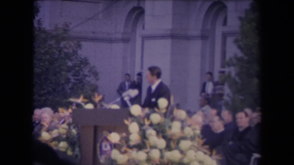 The speaker is seen in profile at the floral podium, with audience members and arched windows behind him.