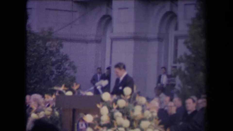 A handheld 8mm frame shows the suited speaker leaning toward the podium, partly obscured by flowers and spectators.