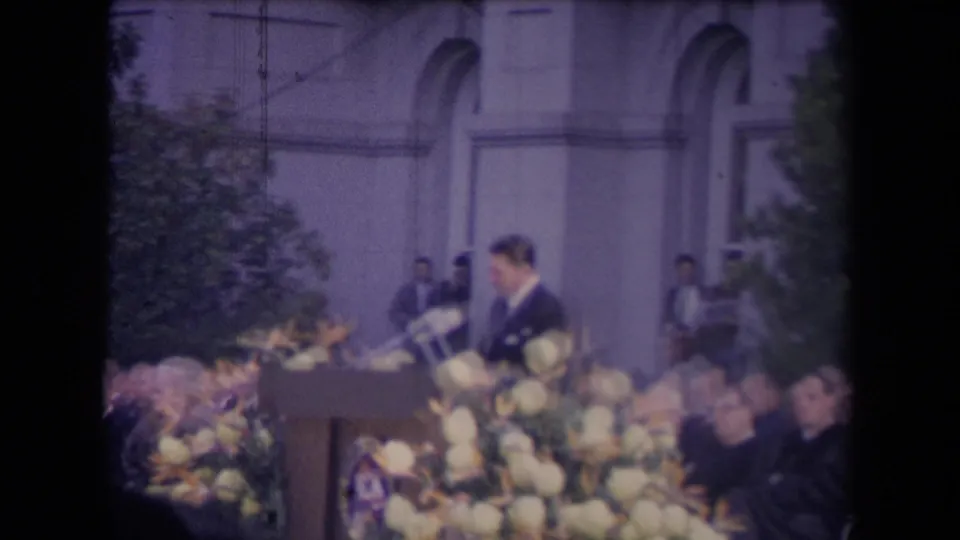 The final frame holds on the formal podium scene, showing flowers, guests, microphones, and the building facade.