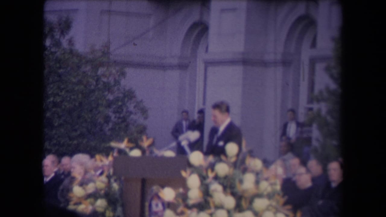 Man in dark suit stands at a flowered podium before a seated crowd.