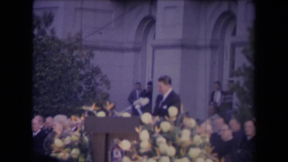 A man in a dark suit stands at a podium with microphones and yellow flowers during an outdoor civic ceremony.