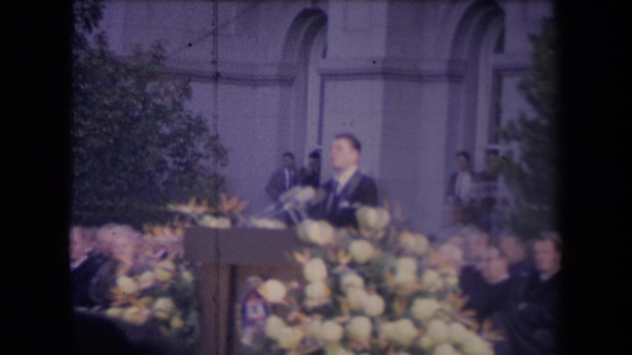 The suited speaker appears in profile at a microphone-covered podium, with attendees and an arched building behind him.