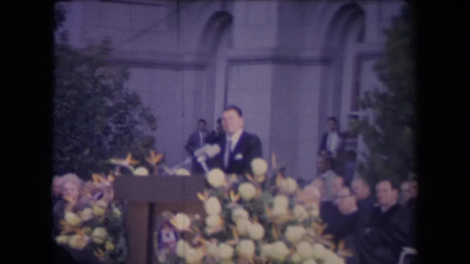 A suited man addresses a formal outdoor crowd from a flowered podium, filmed with soft 8mm color and handheld framing.