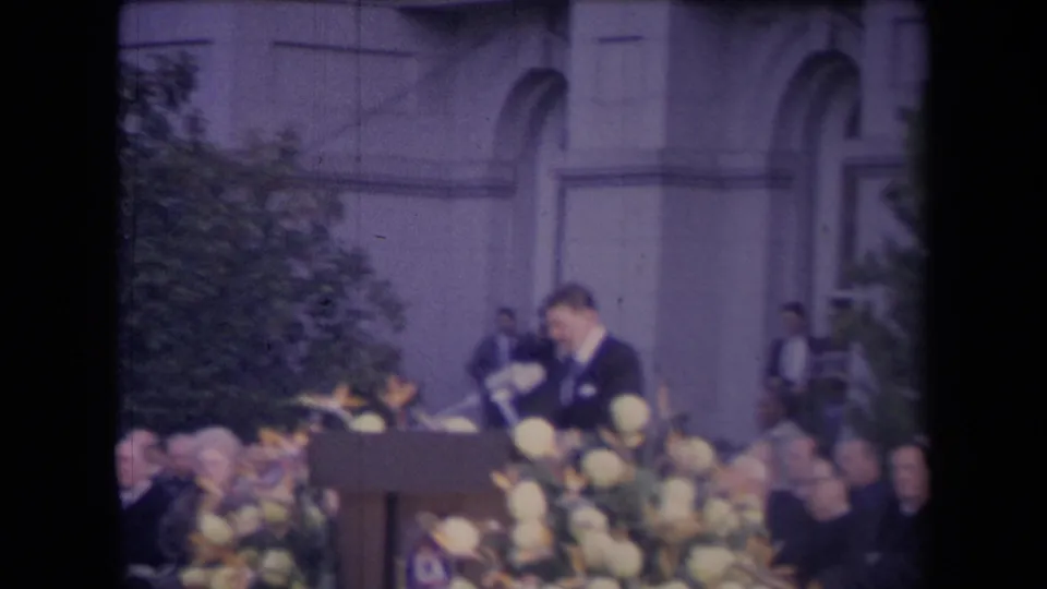 The speaker looks down at the podium or notes, with microphones, flowers, seated attendees, and arched windows in view.