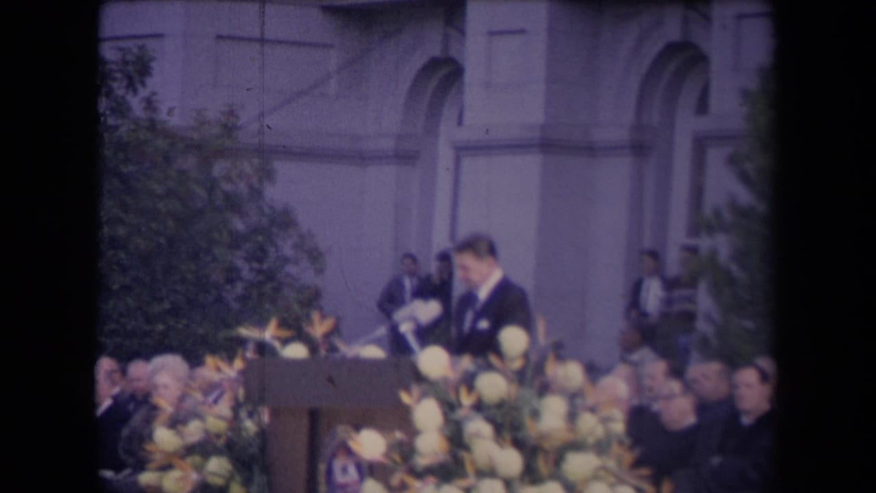 A man at a podium with microphones addresses a seated audience during a formal outdoor ceremony, with arched building details behind him.