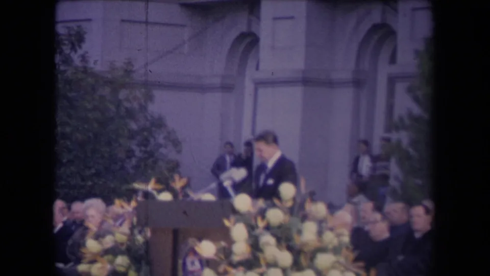 A man at a podium with microphones addresses a seated audience during a formal outdoor ceremony, with arched building details behind him.