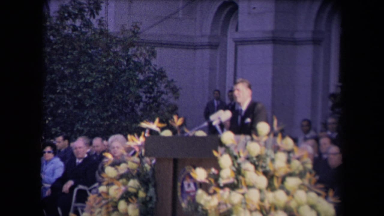 A formally dressed speaker stands at a podium with microphones and flowers while a seated crowd watches outside a classical-style building.