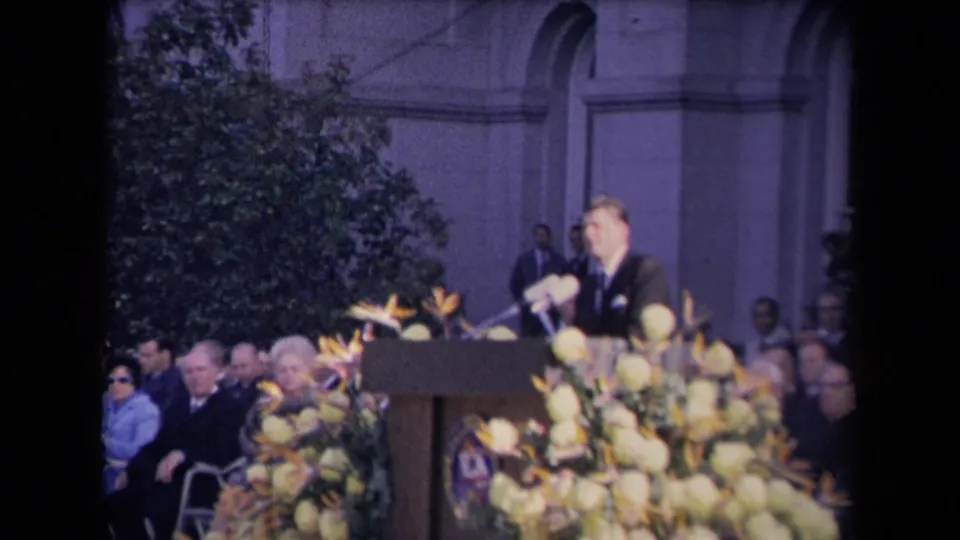 A formally dressed speaker stands at a podium with microphones and flowers while a seated crowd watches outside a classical-style building.