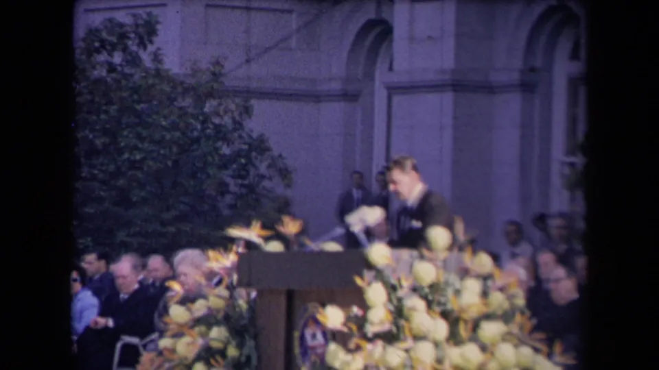 The speaker appears to lean toward the podium microphones, with large flowers in the foreground and attendees seated along the building wall.